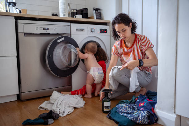 A mom washes baby clothes while her baby curiously plays with the washing machine in a home laundry room.