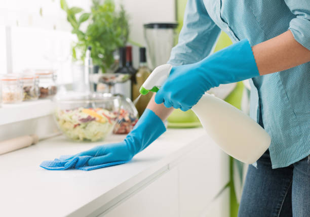 Person in blue gloves cleaning a kitchen counter with a spray bottle and cloth before guests arrive.